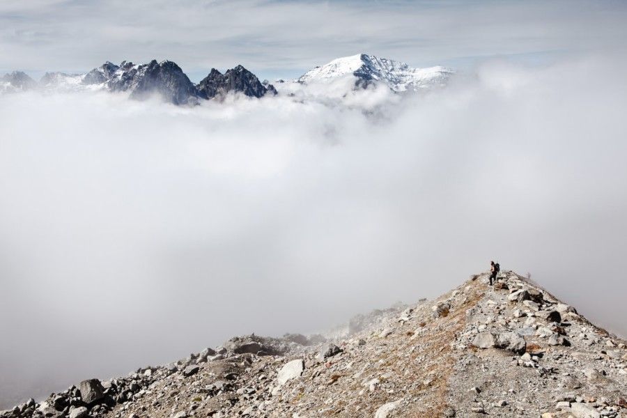 person-standing-on-peak-front-of-white-clouds-and-ice-capped-mountain